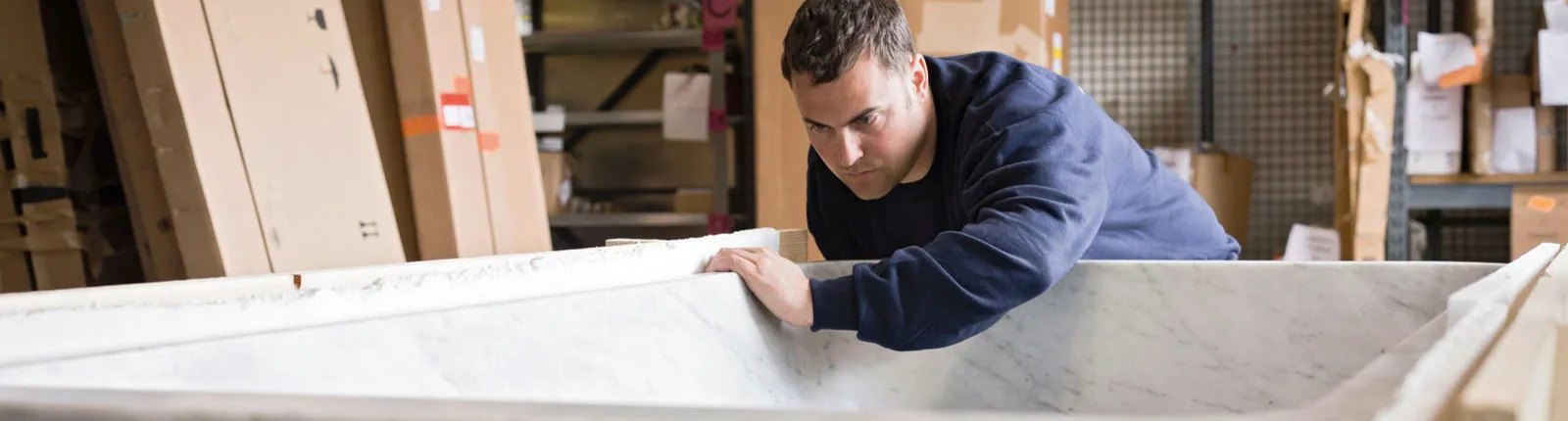 Ein Handwerker inspiziert sorgfältig eine große Badewanne aus Marmor, umgeben von Kartons und Regalen in einer Werkstatt.