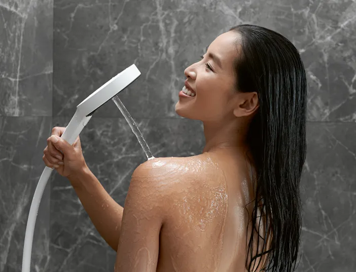 Eine Frau mit langen, nassen Haaren lächelt, während sie eine moderne, weiße Handbrause über ihre Schulter hält und Wasser auf ihren Rücken spritzt.